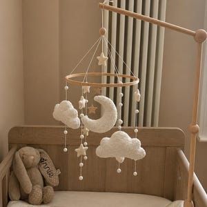 Pode incluir: Móvel de bebé em madeira com uma moldura circular, com nuvens de feltro, estrelas e uma lua crescente. O móvel está pendurado sobre um berço de madeira com um coelhinho de pelúcia. A paleta de cores é neutra, com tons bege e branco.