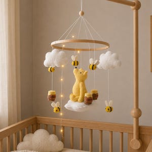 May include: A wooden baby mobile featuring a yellow bear, white clouds, and yellow and black bees. The mobile includes small honey pots and is suspended above a wooden crib. The mobile is lit with small fairy lights.
