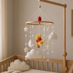 May include: A wooden baby mobile with a Winnie the Pooh character, stars, clouds, and white pom-poms. The mobile is suspended above a wooden crib. The Winnie the Pooh character is yellow and wearing a red shirt.