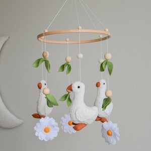 May include: A white felt baby mobile with three white geese holding green leaves, three white felt flowers with orange centers, and wooden beads hanging from a wooden ring.