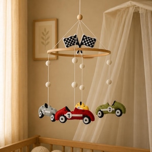 Può includere: Mobile per neonati con quattro macchinine da corsa in feltro grigio, rosso, giallo e verde, ognuna con un numero. Le macchinine sono appese a un anello di legno con bandiere a scacchi e pompon bianchi.