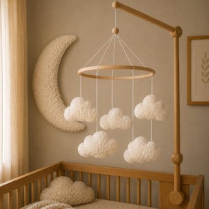 Puede incluir: Móvil para bebé de madera con formas de nubes blancas y esponjosas suspendidas de un marco circular. El móvil cuelga sobre una cuna de madera. Una gran luna creciente texturizada decora la pared. La habitación tiene una paleta de colores neutros.