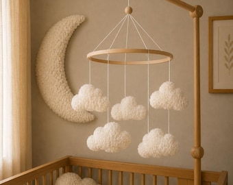 Op de afbeelding: Een houten baby mobile met zachte witte wolken die aan een cirkelvormig frame hangen. De mobile hangt boven een houten wieg. Een grote, getextureerde halve maan siert de muur. De kamer heeft een neutraal kleurenpalet.
