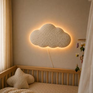 Könnte beinhalten: Eine wolkenförmige Wandleuchte mit weicher Textur und warmem Leuchtkontur. Die Leuchte ist über einem Holzbett montiert. Ein sternförmiges Kissen und ein Mobile mit Filzdekorationen sind ebenfalls zu sehen.