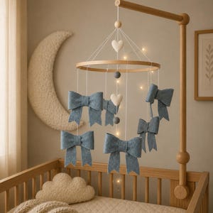 May include: A baby mobile featuring six blue felt bows, two white felt hearts, and two grey felt balls, suspended above a wooden cot. The cot has a beige quilted mattress and a cloud-shaped pillow. Fairy lights are strung around the mobile.