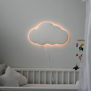 Pode incluir: Uma luz noturna em forma de nuvem branca com um brilho quente ilumina um berço branco com um travesseiro em forma de estrela branca. Um móbile branco com flores rosa e amarelas e abelhas pendura sobre o berço.