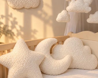 Könnte beinhalten: Ein Set aus cremefarbenen, strukturierten Plüschkissen in Form eines Sterns, einer Mondsichel und einer Wolke, angeordnet in einem Holzbettchen. Ein passendes Mobile mit hängenden Wolken und einem kleinen braunen Bären hängt darüber.