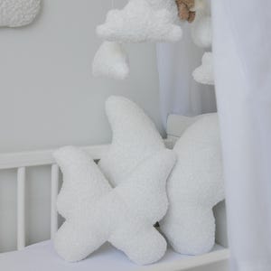 Puede incluir: Cuna blanca con un m&oacute;vil de nubes blancas y esponjosas colgando sobre ella. Dos almohadas blancas en forma de nube esponjosa est&aacute;n en el colch&oacute;n de la cuna.