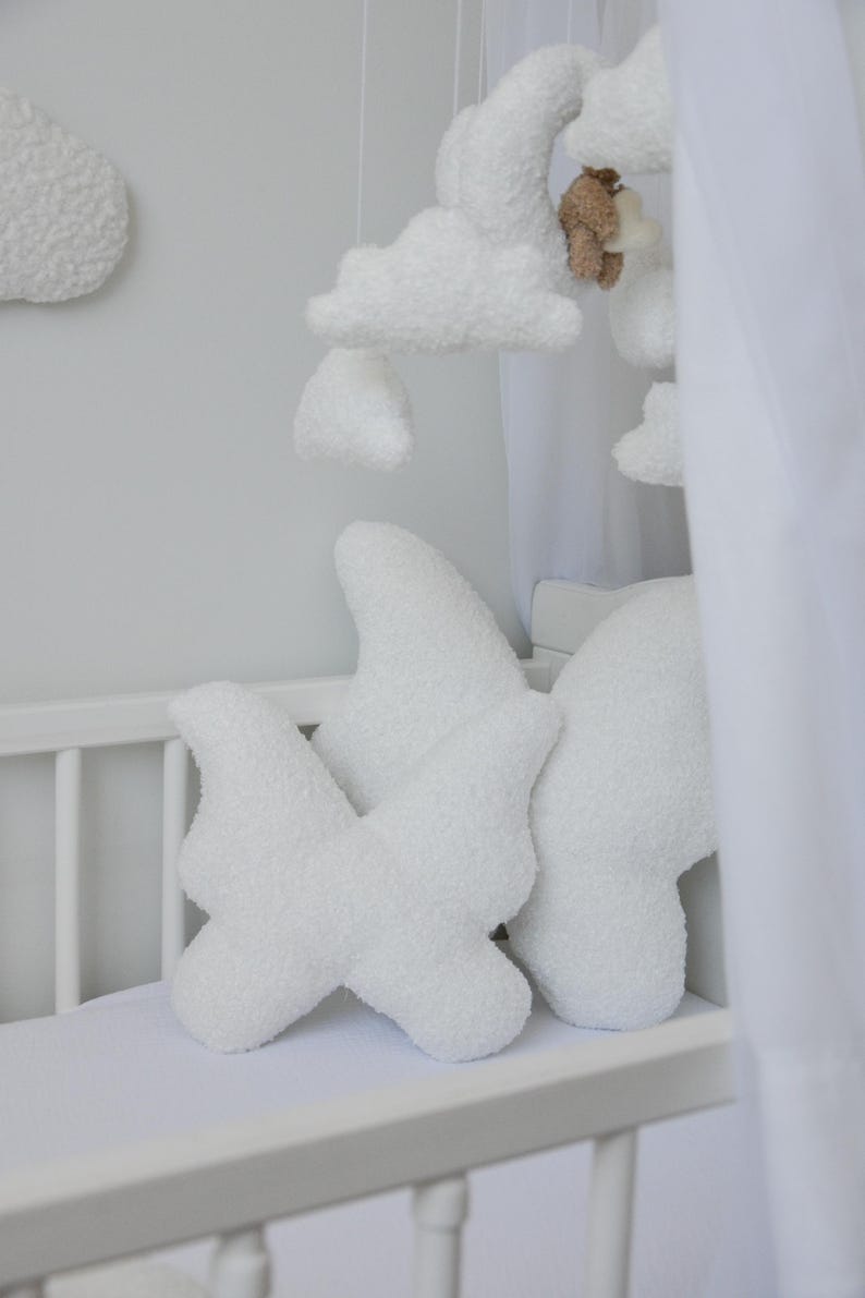 Puede incluir: Cuna blanca con un m&oacute;vil de nubes blancas y esponjosas colgando sobre ella. Dos almohadas blancas en forma de nube esponjosa est&aacute;n en el colch&oacute;n de la cuna.