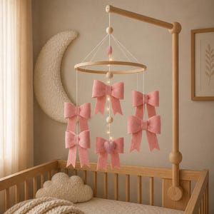 May include: A baby mobile made of wood, featuring six pink felt bows hanging from a circular frame. The mobile is positioned above a wooden cot. The cot contains a beige blanket and a cloud-shaped pillow.