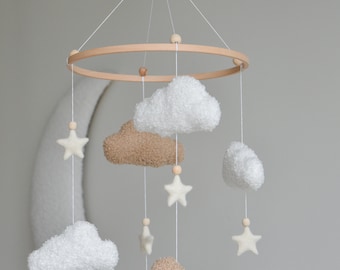 Op de afbeelding: Een houten babymobiel met witte en bruine pluche wolken en witte viltsterren die aan een houten ring hangen. De mobiel is aan het plafond gehangen met wit touw.