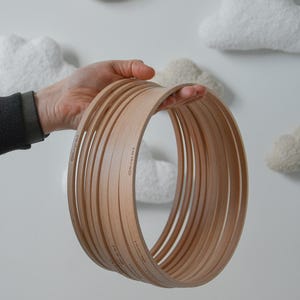 Puede incluir: Una pila de aros de bordado de madera marrón claro sostenidos en una mano. Los aros están apilados, mostrando su forma circular. El fondo incluye nubes blancas y una luna creciente.