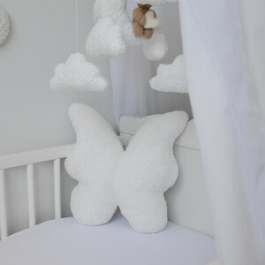 Puede incluir: Cuna blanca con una almohada con forma de mariposa blanca y un dosel blanco. La cuna tiene un m&oacute;vil blanco colgado encima con nubes y un oso.