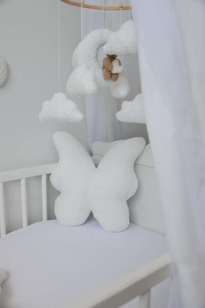 Puede incluir: Cuna blanca con una almohada con forma de mariposa blanca y un dosel blanco. La cuna tiene un m&oacute;vil blanco colgado encima con nubes y un oso.