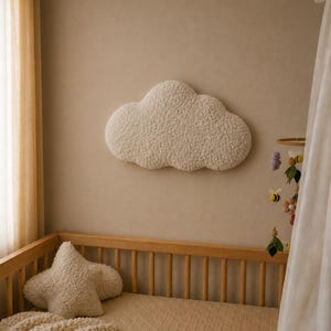 Könnte beinhalten: Eine flauschige, cremefarbene, wolkenförmige Wanddekoration hängt über einem Holzbettchen. Ein sternförmiges Kissen und eine gesteppte Decke befinden sich im Bettchen. Ein Mobile mit Bienen und Blumen hängt in der Nähe.