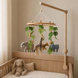 May include: A wooden baby mobile featuring felt animal figures of a giraffe, leopard, elephant, and rhino, with green leaf accents. The mobile hangs above a wooden crib with a stuffed bunny and pillow.