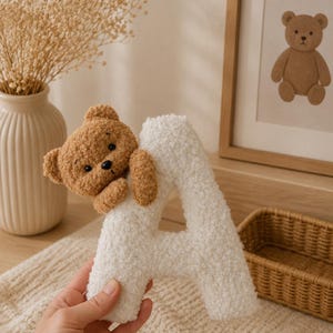 May include: A fluffy white letter 'A' with a brown teddy bear attached. The bear has a stitched face and is hugging the letter. A framed bear illustration and a woven basket are in the background.