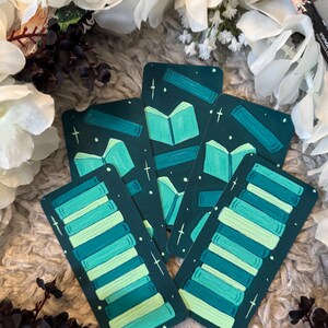 May include: A collection of teal and green book-themed bookmarks. Each bookmark features a stack of books or an open book design with white star accents on a dark teal background. The bookmarks are arranged on a soft, textured surface.