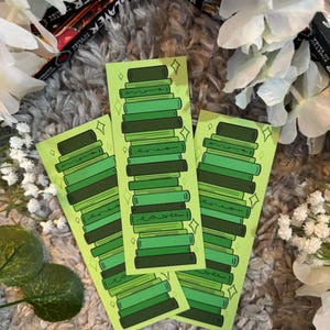 Könnte beinhalten: Drei grüne Lesezeichen mit einem Bücherstapel-Design. Die Lesezeichen zeigen verschiedene Grüntöne von Büchern mit einem hellgrünen Hintergrund und funkelnden Akzenten. Die Lesezeichen sind auf einer grauen, strukturierten Oberfläche mit weißen Blumen und Büchern im Hintergrund angeordnet.