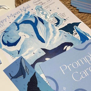 May include: An assortment of blue and white ocean-themed stationery, including stickers, cards, and a newsletter. The newsletter features illustrations of whales, jellyfish, and other marine life. The text "Happy Moon Mail" is visible.