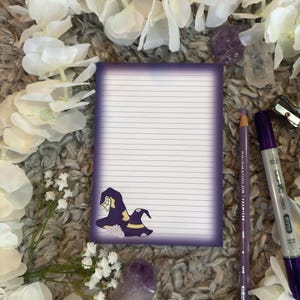 May include: Purple lined notepad with a witch hat illustration. The notepad is on a fuzzy brown surface with white flowers and purple crystals.