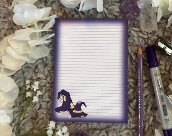 May include: Purple lined notepad with a witch hat illustration. The notepad is on a fuzzy brown surface with white flowers and purple crystals.