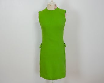 Puede incluir: Un vestido sin mangas de color verde lima con una silueta recta. El vestido presenta pequeños lazos decorativos a cada lado cerca de la cintura. El vestido se exhibe en un maniquí sobre un fondo blanco liso.