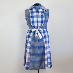 Könnte beinhalten: Ein ärmelloses blau-weiß kariertes Kleid mit weißem Bindegürtel. Das Kleid wird auf einer Schaufensterpuppe vor weißem Hintergrund präsentiert. Es hat einen Kragen und eine Knopfleiste.