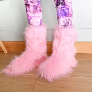 May include: A pair of pink faux fur boots. The boots have a fluffy texture and are worn with leggings featuring a floral rose pattern in shades of purple and pink. The boots are on a wooden floor.