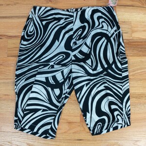 Könnte beinhalten: Schwarz-weiß gemusterte Caprihose. Die Caprihose hat ein wirbelndes, abstraktes Design. Der Stoff ist hell mit schwarzen Linien. Die Hose liegt gefaltet auf einer Holzoberfläche.
