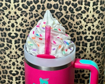May include: A pink Stanley travel mug with a turquoise handle and a white lid. The mug has a logo that says "STANLEY" in turquoise lettering. The mug is filled with whipped cream and colorful sprinkles. A pink straw is sticking out of the whipped cream.