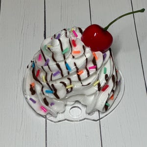 May include: A white cupcake with chocolate frosting and colorful sprinkles topped with a red cherry. The cupcake is on a clear plastic base.