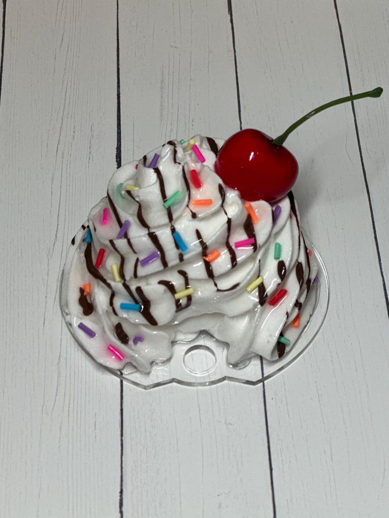 May include: A white cupcake with chocolate frosting and colorful sprinkles topped with a red cherry. The cupcake is on a clear plastic base.