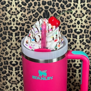 May include: A pink Stanley travel mug with a teal handle and a white lid. The mug has a teal logo that says "STANLEY". The lid is topped with whipped cream, chocolate syrup, sprinkles, and a red cherry.