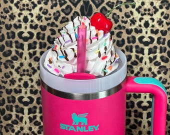 May include: A pink Stanley travel mug with a teal handle and a white lid. The mug has a teal logo that says "STANLEY". The lid is topped with whipped cream, chocolate syrup, sprinkles, and a red cherry.