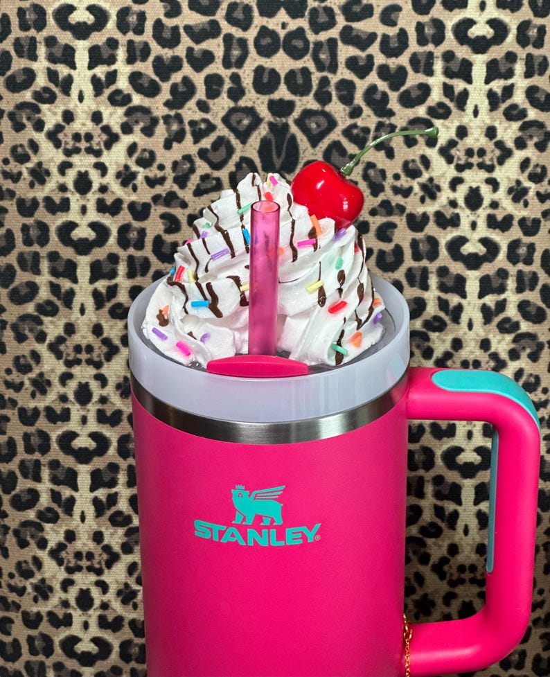 May include: A pink Stanley travel mug with a teal handle and a white lid. The mug has a teal logo that says "STANLEY". The lid is topped with whipped cream, chocolate syrup, sprinkles, and a red cherry.