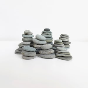 Puede incluir: Varias pilas de piedras de río grises lisas de diferentes tamaños están dispuestas sobre una superficie blanca. Las piedras son en tonos de gris y gris azulado, creando una estética natural y minimalista. Las pilas varían en altura y forma.