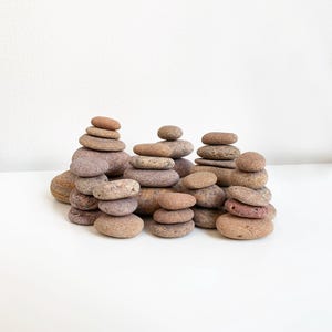 Puede incluir: Una colección de pequeñas piedras lisas, marrones y grises, apiladas a diferentes alturas sobre una superficie blanca. Las piedras están dispuestas de forma aleatoria, creando una escena visualmente atractiva y relajante.