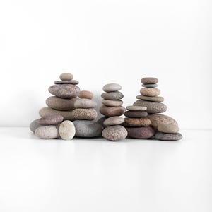 Puede incluir: Una colección de piedras grises, marrones y tostadas apiladas en una formación equilibrada sobre una superficie blanca. Las piedras están dispuestas en tres grupos, con el grupo más alto en el centro.