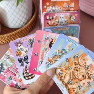 May include: A collection of rectangular savings challenge cards featuring colourful illustrations of pumpkins, ghosts, and animals. Each card displays various monetary values in British pounds, such as £1, £2, £3, £4, and £5, alongside themed titles like Happy Little Harvest, Spooky Magic, and Highland.