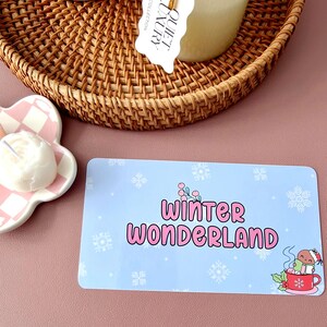 May include: A light blue rectangular card with the words "Winter Wonderland" in pink, surrounded by snowflakes. A cartoon character in a Santa hat is in a red mug. A woven basket and a candle are in the background.