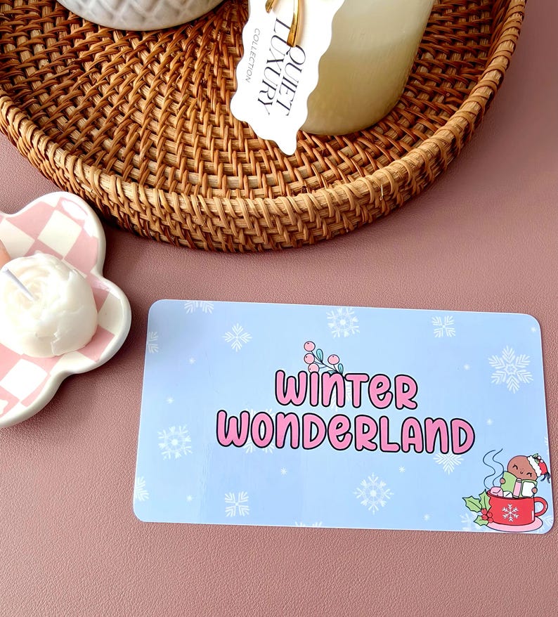 May include: A light blue rectangular card with the words "Winter Wonderland" in pink, surrounded by snowflakes. A cartoon character in a Santa hat is in a red mug. A woven basket and a candle are in the background.