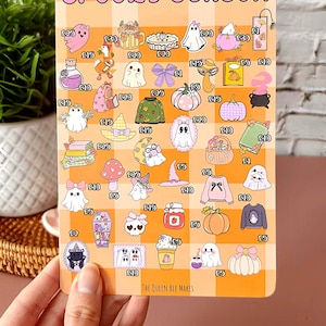 May include: A rectangular sticker sheet with a checkered orange background and the words "SPOOKY SEASON" at the top. The sheet features various Halloween-themed illustrations, including ghosts, pumpkins, and sweaters. The Queen Bee Makes is printed at the bottom.