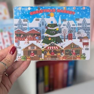 May include: A Christmas market game board with a blue background and white snow. The board features a Christmas tree, various stalls, and buildings. The stalls have prices listed in pounds sterling. The text "Christmas Market" is at the top of the board. The text "The Queen Bee Makes" is at the bottom of the board.