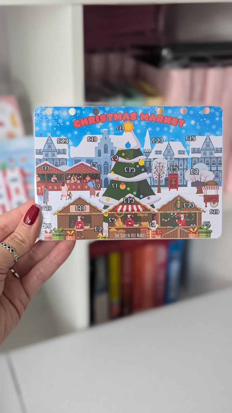 May include: A Christmas market game board with a blue background and white snow. The board features a Christmas tree, various stalls, and buildings. The stalls have prices listed in pounds sterling. The text "Christmas Market" is at the top of the board. The text "The Queen Bee Makes" is at the bottom of the board.