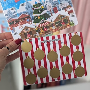 May include: A Christmas market themed savings challenge card with a red and white striped background and gold circles. The text "Merry Mystery Saving Challenge" is printed on the card.