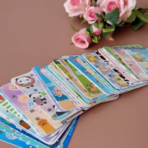 May include: A pile of colourful cards featuring cartoon animal illustrations, such as pandas, ducks, and octopuses. The cards have a blue border and are stacked on a pink surface. Pink roses are in the background.