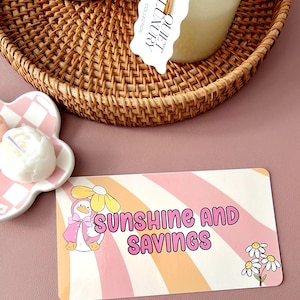 May include: A woven wicker tray holds a candle and a ceramic container. A card with the text "SUNSHINE AND SAVINGS" features a cartoon duck wearing a pink cape and a daisy. A small, flower-shaped dish holds a white candle.
