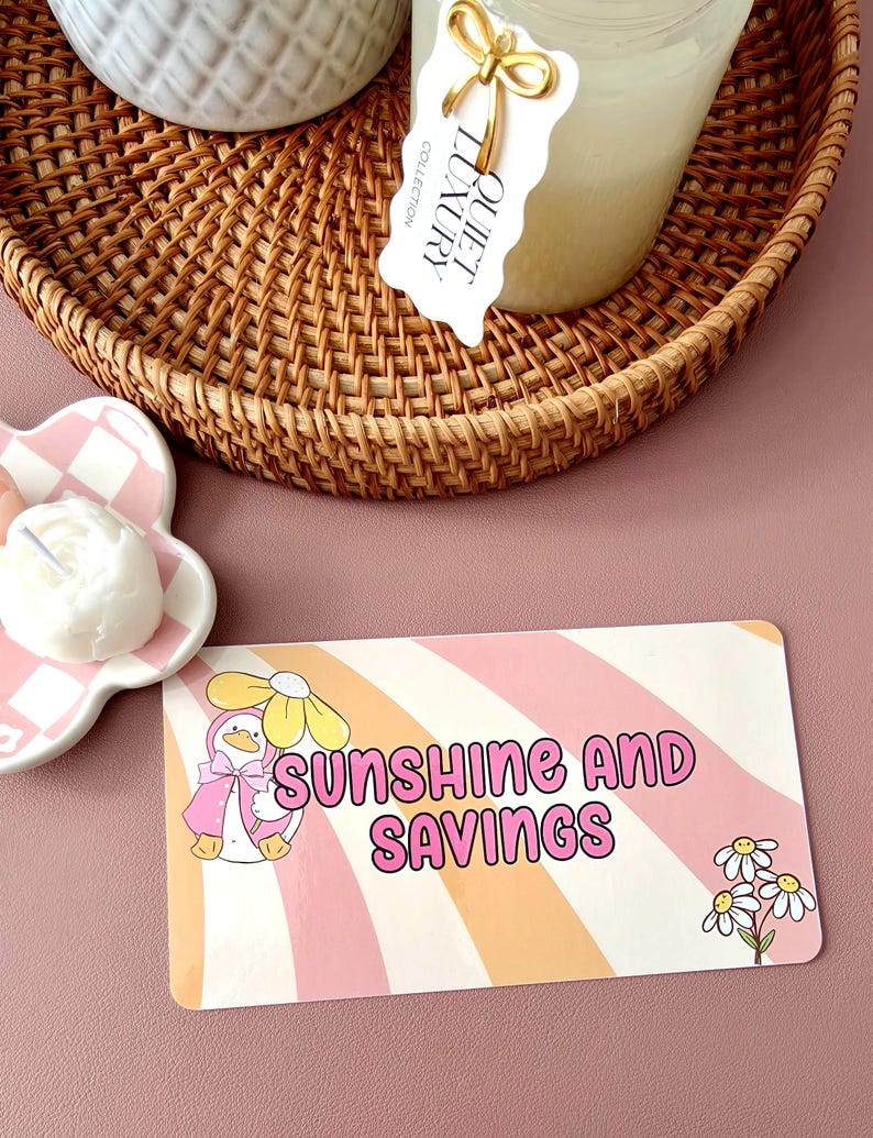 May include: A woven wicker tray holds a candle and a ceramic container. A card with the text "SUNSHINE AND SAVINGS" features a cartoon duck wearing a pink cape and a daisy. A small, flower-shaped dish holds a white candle.