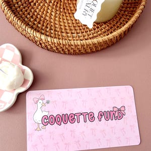 May include: A pink rectangular card with the words "COQUETTE FUND" in pink bubble letters, and a cartoon goose with a pink bow. The background has a pattern of pink bows. A woven basket and a candle are in the background.